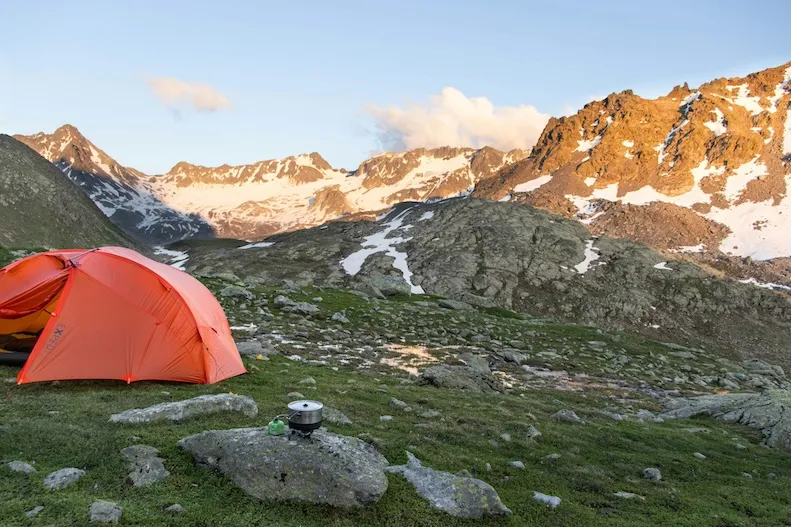 An orange tent pitched on alpine tundra at sunrise, snow-streaked peaks behind it and a camp stove boiling water on a nearby rock.