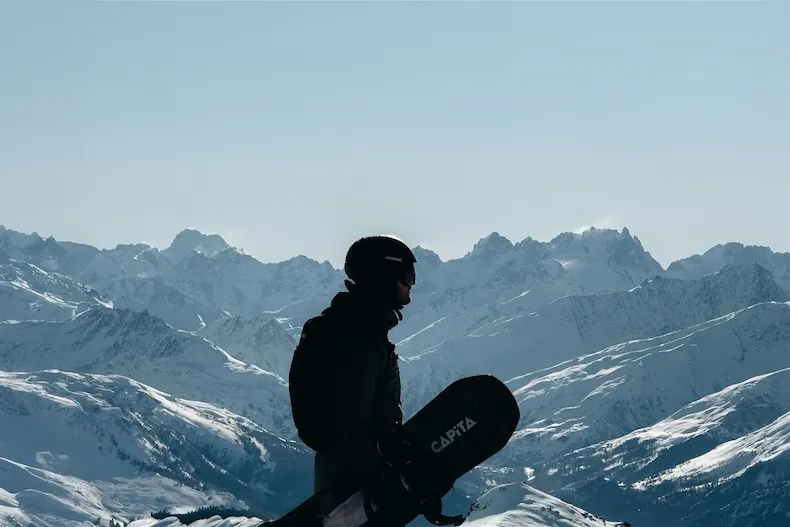 The silhouette of a snowboarder holding their board, backed by a wide range of snow-covered peaks.
